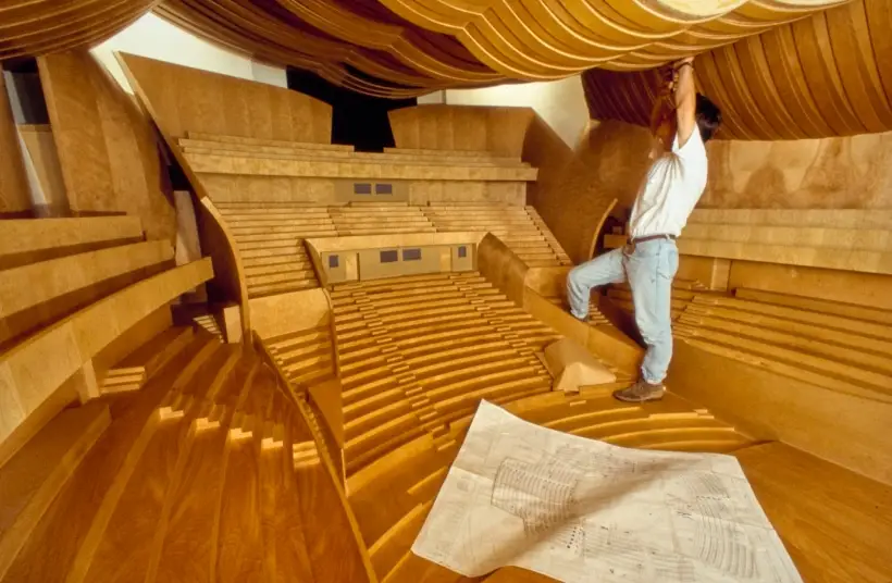 A person standing inside a large, scale model of the Walt Disney Concert Hall interior makes adjustments to the model. A plan drawing lies on the floor in front of them.