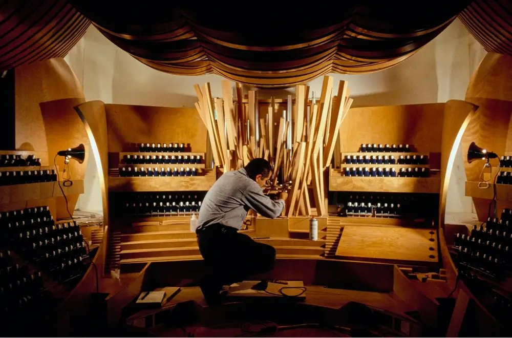 A person kneeling inside a large, scale model of the Walt Disney Concert Hall interior adjusts the organ at its center.