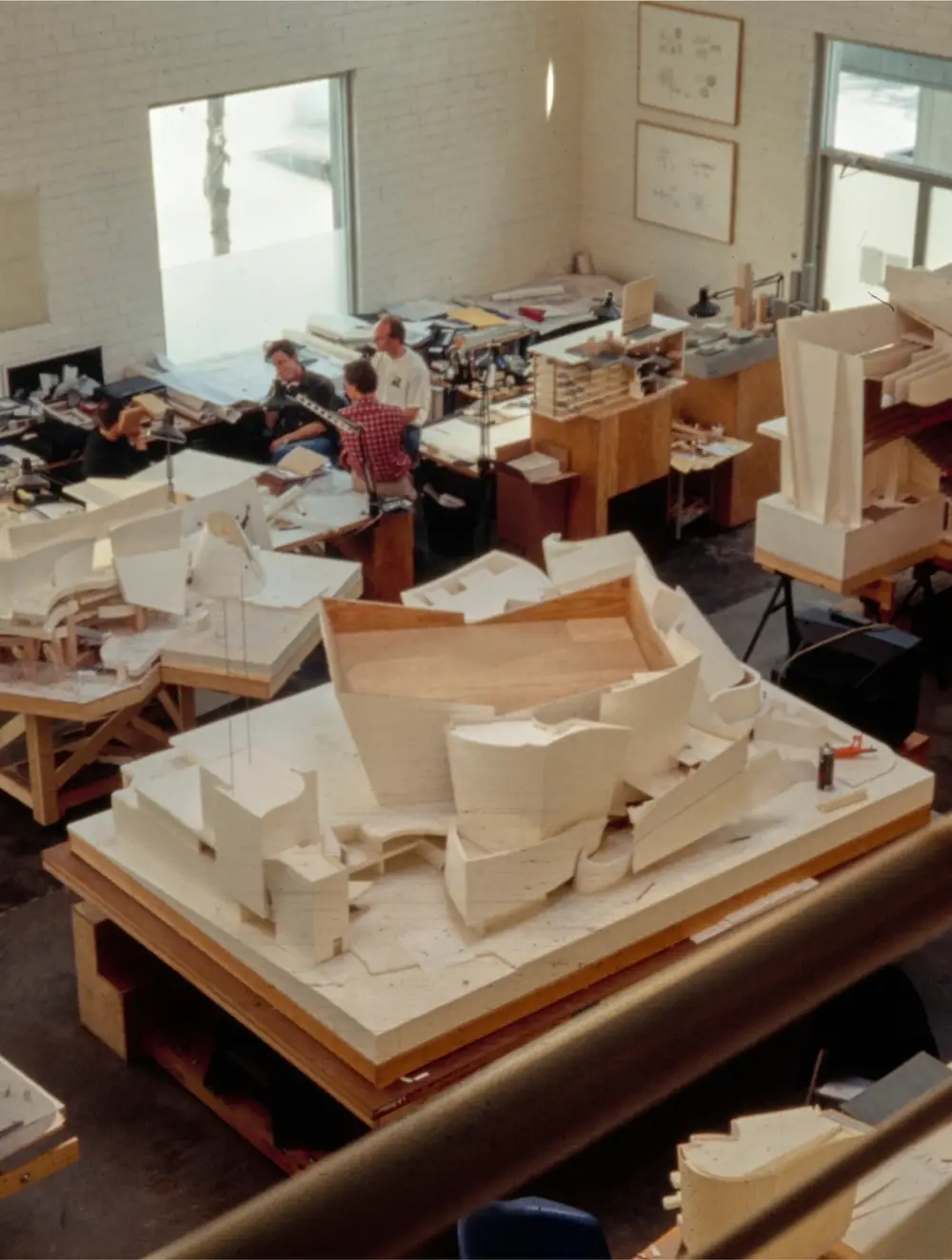 An architect's studio is filled with models of Walt Disney Concert Hall, both large and small. Three people stand nearby, examining one of these models.