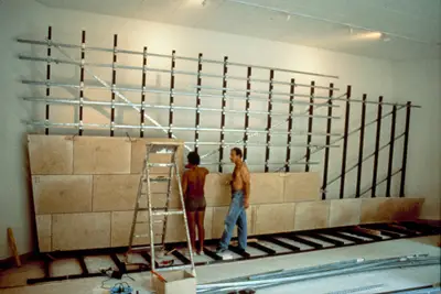Two shirtless workers in a non-descript office affix stone panels to a metal frame. Around them, construction materials, tools, and a ladder suggest the work still left to be done.