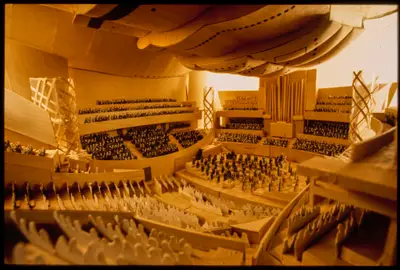 A view into an interior model of Walt Disney Concert Hall roughly suggests the appearance of a performance, with audience members in seats and performers on stage both represented with cutout images. The wooden interior is awash with artificial light.