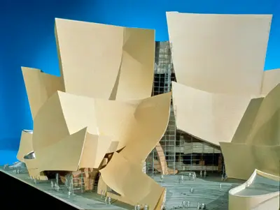 A dramatically lit model of Walt Disney Concert Hall clad in beige textured paper sits against a blue background, highlighting the building's curving forms. Figurines and model cars sit in the foreground.
