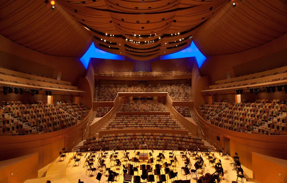 An interior view of Walt Disney Concert Hall from behind the stage looking out toward the hall. The hall and seats that surround the stage on all sides are empty, showcasing the sweeping forms and warm colors of the hall's interior architecture.