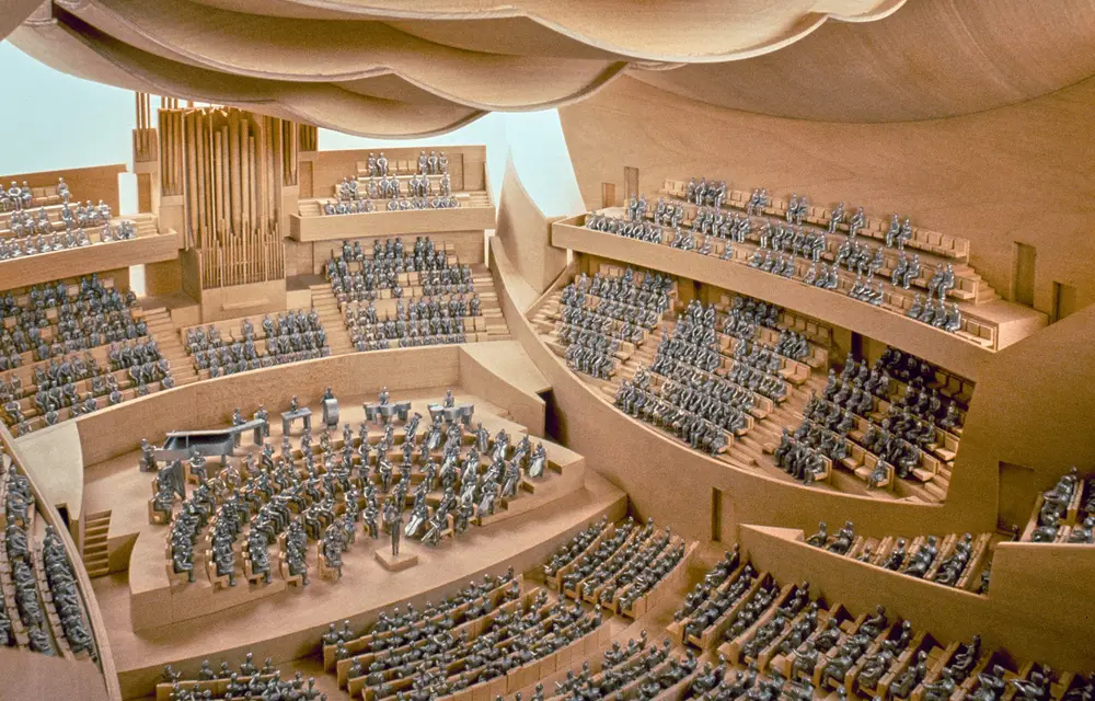 A view into a highly detailed interior model of Walt Disney Concert Hall suggests the appearance of a performance, with audience members in seats and performers on stage both represented in silver figurines. The wooden interior is awash with artificial light.