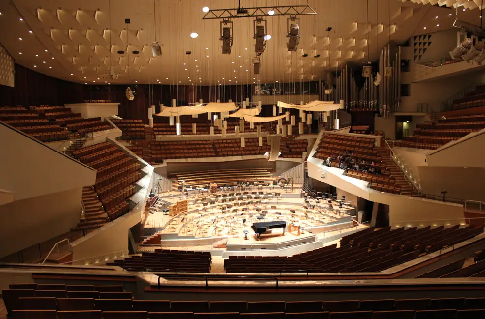 An interior view of the Berlin Philharmonie's Grand Hall from an upper level towards the stage, which is surrounded on all sides by seats. A single performer sits at a piano in the center of the stage while the rest of the hall is empty, showcasing the hall's geometry and warm colors.