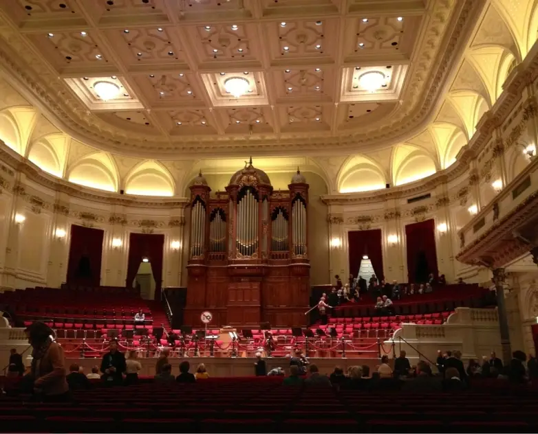 An interior view of the Main Hall of the Concertgebouw, Amsterdam from an upper level towards the stage and organ during a performance. The hall is filled with natural light and the audience is seated in front of the stage and in galleries on all sides of the stage.
