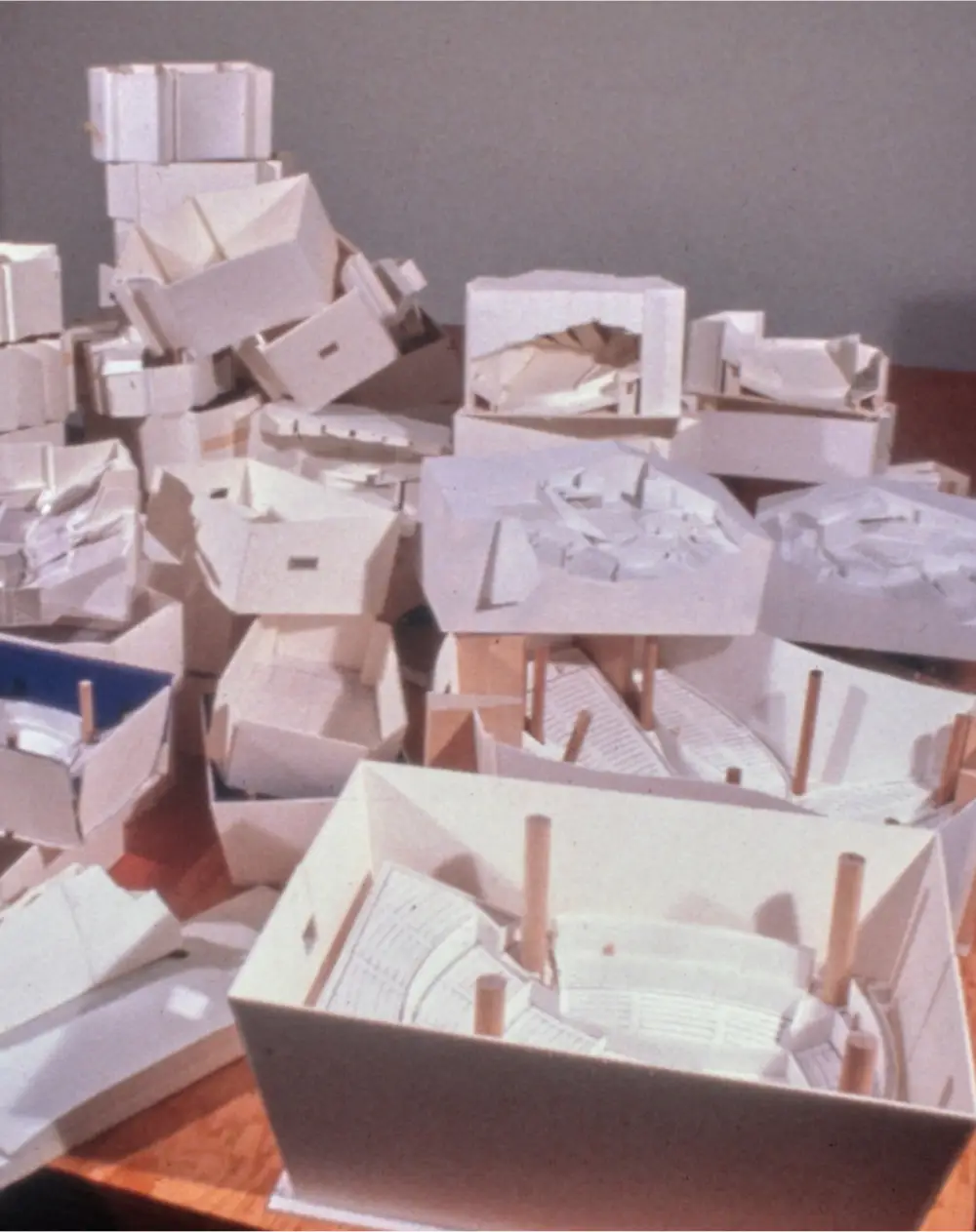 Models in paper and wood accumulate on a desk, piled atop one another.