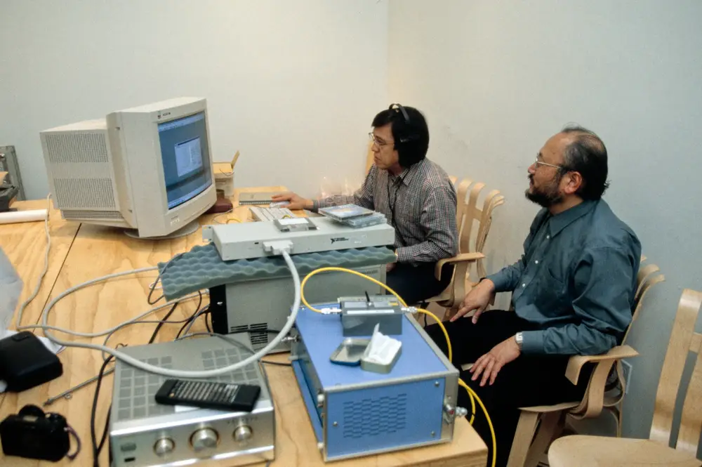 Two people sit in front of a model of a computer and various sound devices, analyzing their data.