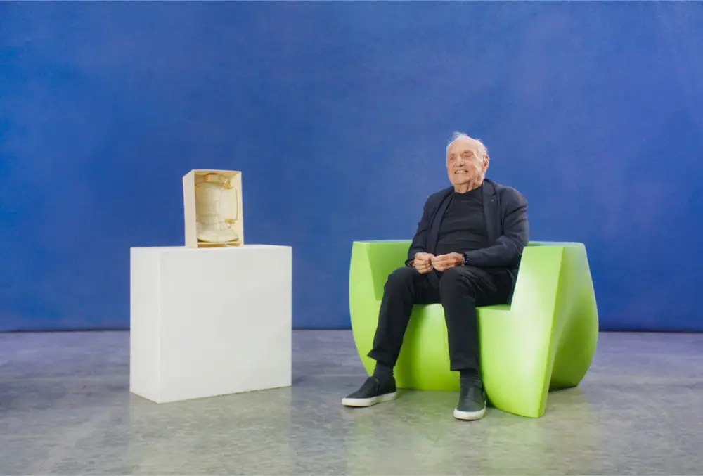 Frank Gehry sits on a bright green chair next to a small study model for the Walt Disney Concert Hall interior, which sits on a white pedestal.