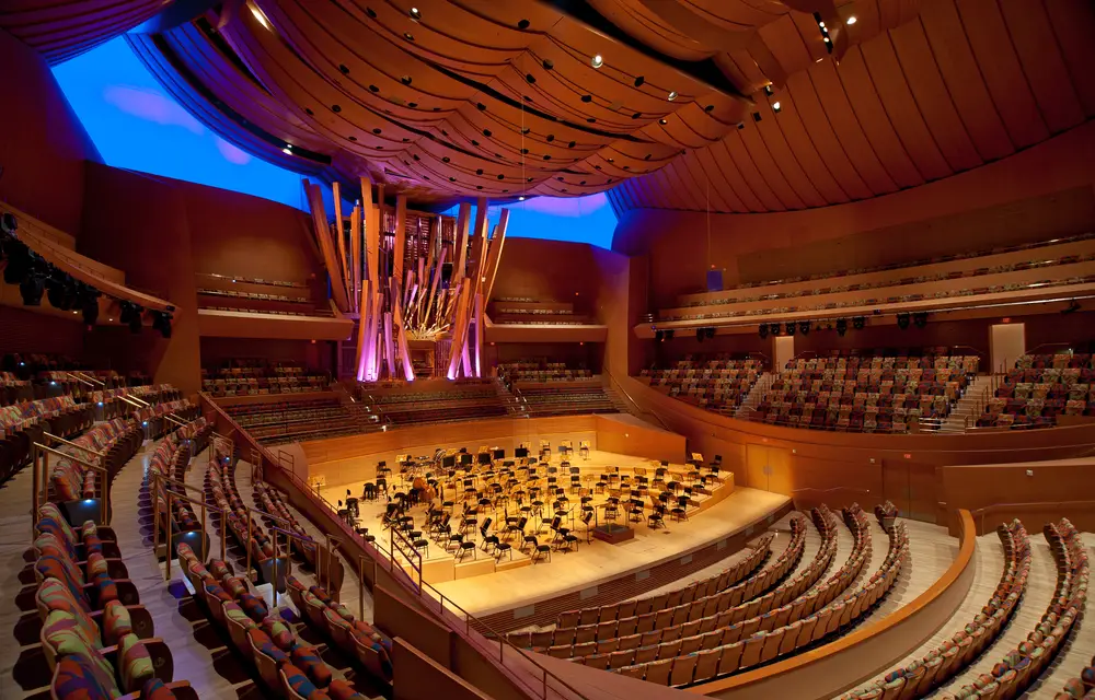 An interior view of Walt Disney Concert Hall from an upper level toward the stage and organ. The hall and seats that surround the stage on all sides are empty, showcasing the sweeping forms and warm colors of the hall's interior architecture.