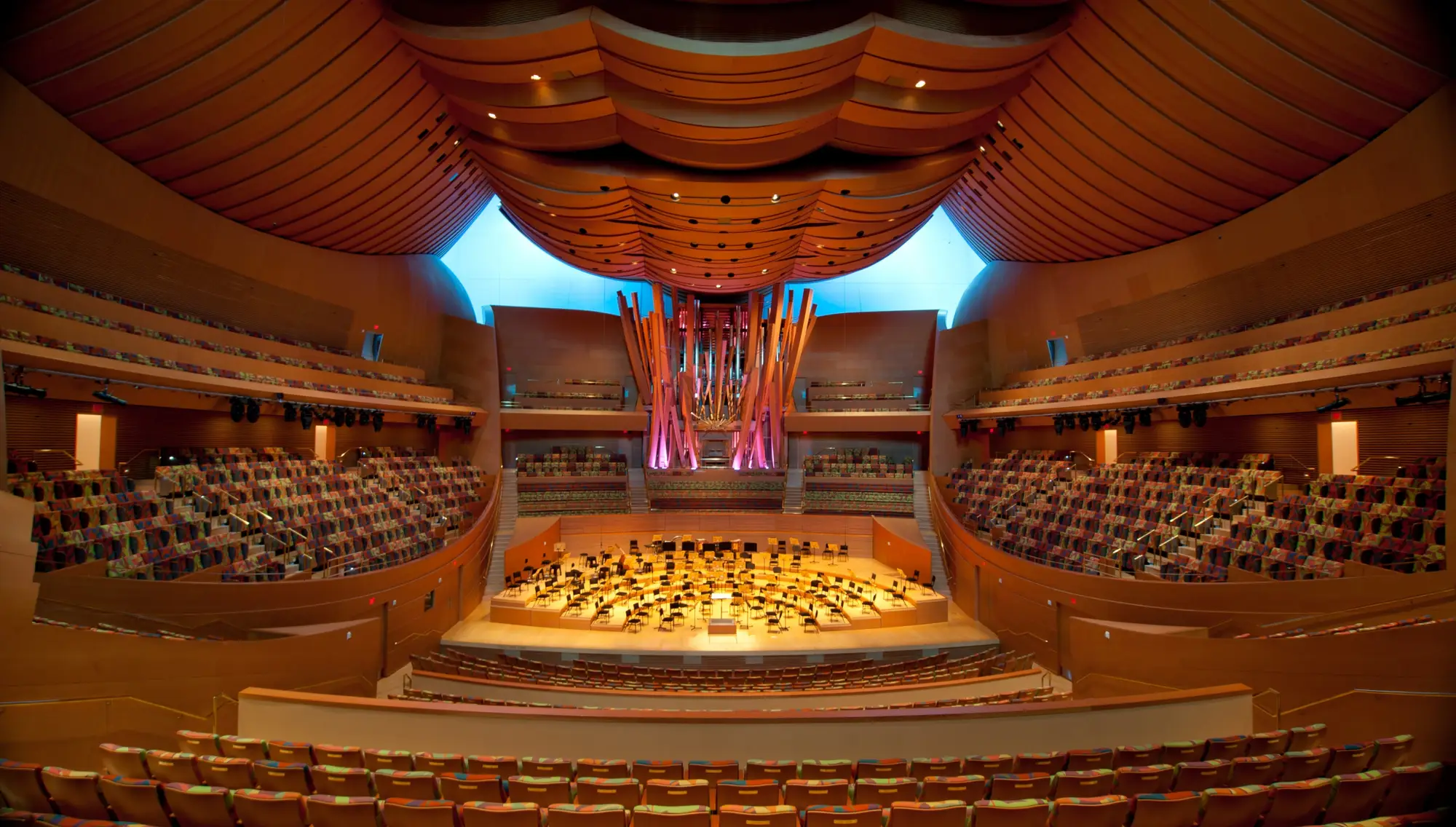 A view of the organ from the stage of Walt Disney Concert Hall. The organ is lit from within by golden light that showcases its rows of internal pipes and is also lit externally by blue light that highlights the upward curves of the organ's largest pipes.
