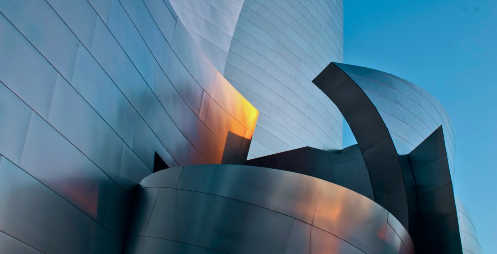 An exterior view of Walt Disney Concert Hall showing the building's complex curves and metal surface reflecting the bright blue sky above it.