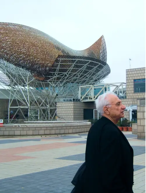 Frank Gehry stands in front of a canopy structure, known as "El Peix [The Fish]," in front of the Hotel Arts Barcelona. The canopy is made up of a latticework of reflective stainless steel that is evocative of a golden swimming fish.