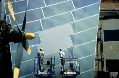 Two construction workers on lifts affix a large metal panel to a wall with many other panels already adhered. A wind turbine sits in the foreground.