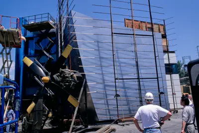 Two construction workers examine a metal wall surrounded by water-spraying equipment, a wind turbine, construction lifts, and other workers milling about.