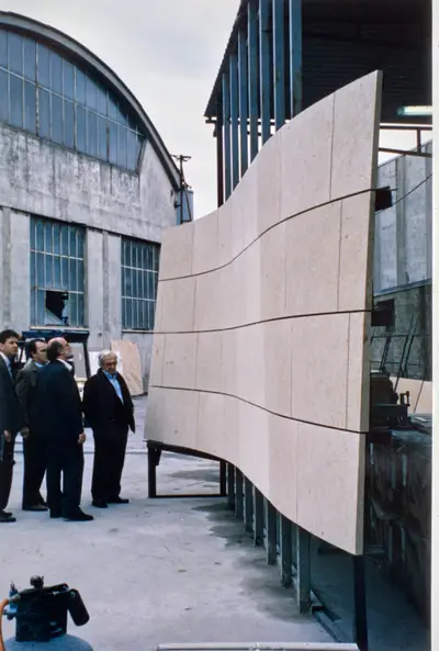 A group of men gathered in an industrial setting closely examine a curving stone wall suspended from a metal frame.