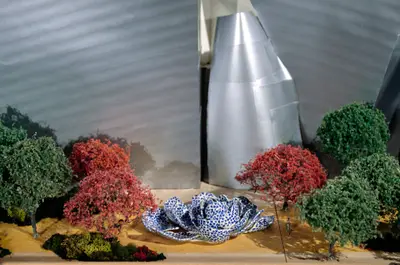 A close-up view of a model of Walt Disney Concert Hall clad in metal-foil paper appears to represent a completed building. In the foreground are bushes and trees with green and red leaves, and at the center is a model of a fountain which takes the form of a rose clad in white and blue tiles.