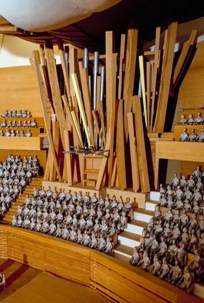 A scale model of an organ constructed in wood with pipes dynamically splayed out in multiple directions. The instrument is placed within a model of the interior of the Walt Disney Concert Hall, with small figurines representing audience members in surrounding seats.