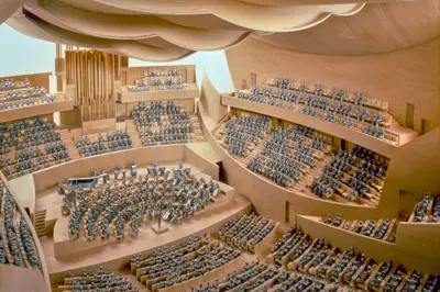 A view into a highly detailed interior model of Walt Disney Concert Hall suggests the appearance of a performance, with audience members in seats and performers on stage both represented in silver figurines. The wooden interior is awash with artificial light.