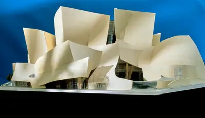A dramatically lit, eye-level view of a model of Walt Disney Concert Hall constructed in textured beige paper, acrylic, and wood against a bright blue background suggests the dynamic appearance of the building.