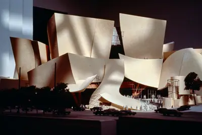 A dramatically lit, eye-level view of a model of Walt Disney Concert Hall constructed in textured beige paper and wood moodily suggests the dynamic appearance of the building. Shadows and scale cars, trees, and figurines afford realism.