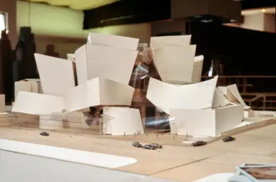 An eye-level view of a model of Walt Disney Concert Hall constructed in wood, acrylic, and paper suggests the dynamic form of the building. Scale cars and figurines afford realism, while polaroids and other office ephemera sit in the foreground and background of the scene.