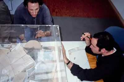 A model of Walt Disney Concert Hall is encased in a clear acrylic box. A top-down view shows two people tracing the model's outlines in red ink on the acrylic.
