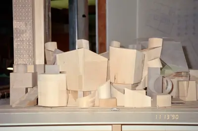 An eye-level view of a model of Walt Disney Concert Hall constructed in wood and acrylic suggests the appearance of a building composed of stacked wooden blocks. Drawings and other office ephemera are visible in the background.