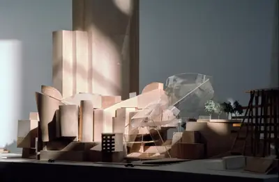 A dramatically lit, eye-level view of a model of Walt Disney Concert Hall constructed in wood and acrylic suggests the appearance of a building composed of vertically oriented wooden blocks, with a wooden tower in the background. Scale cars and trees afford realism.