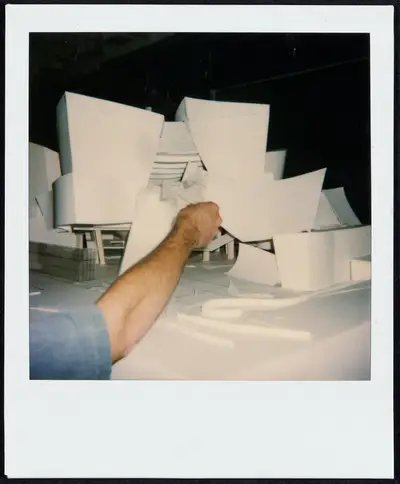 A person's arm reaches to construct a model of the Walt Disney Concert Hall exterior in paper, acrylic, and tape.
